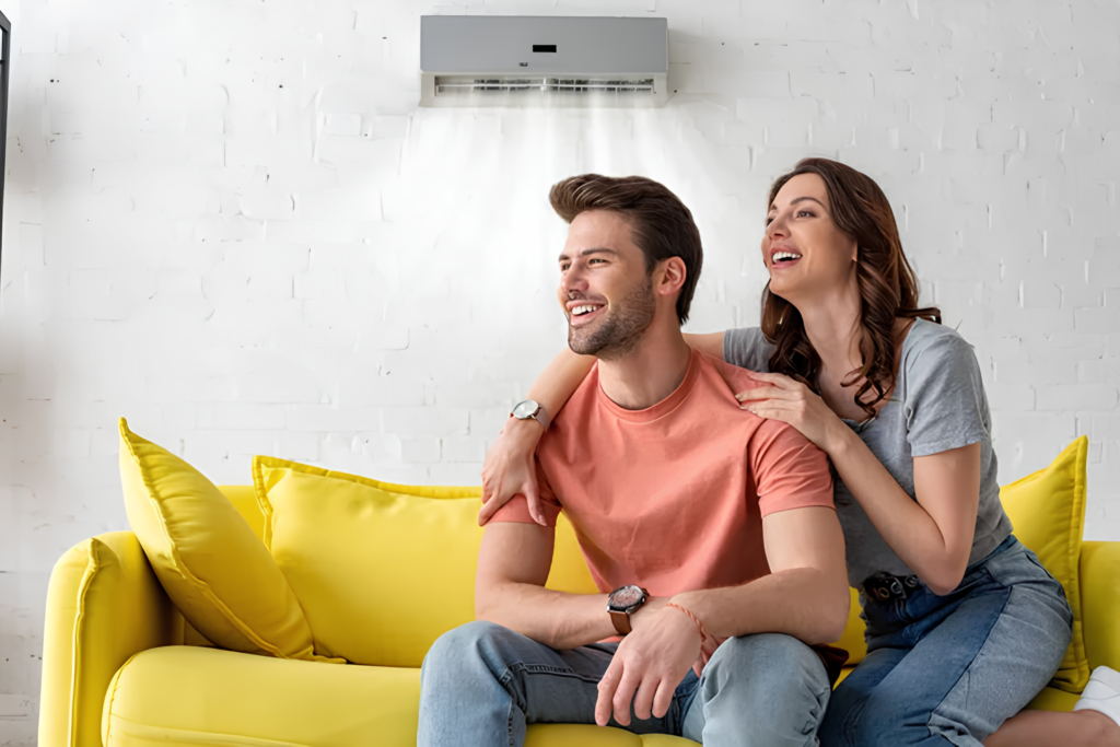Happy couple sitting under an AC unit blowing cool air, representing the cooling and air conditioning services offered by JCC Heating and Cooling.